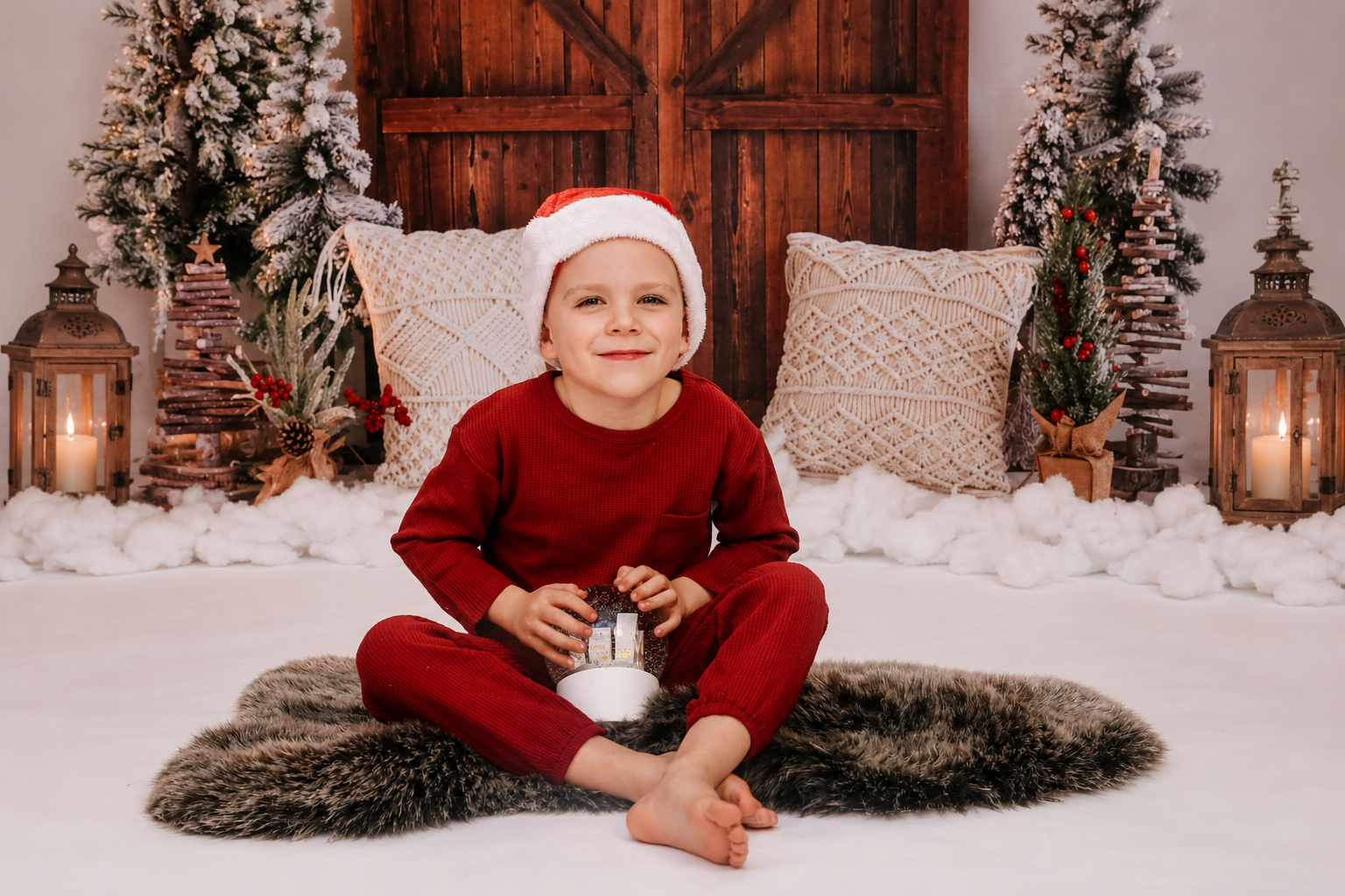 Young boy in red holiday pajamas sitting in a cozy winter portrait scene with snowy decor, wooden doors, and festive studio details.