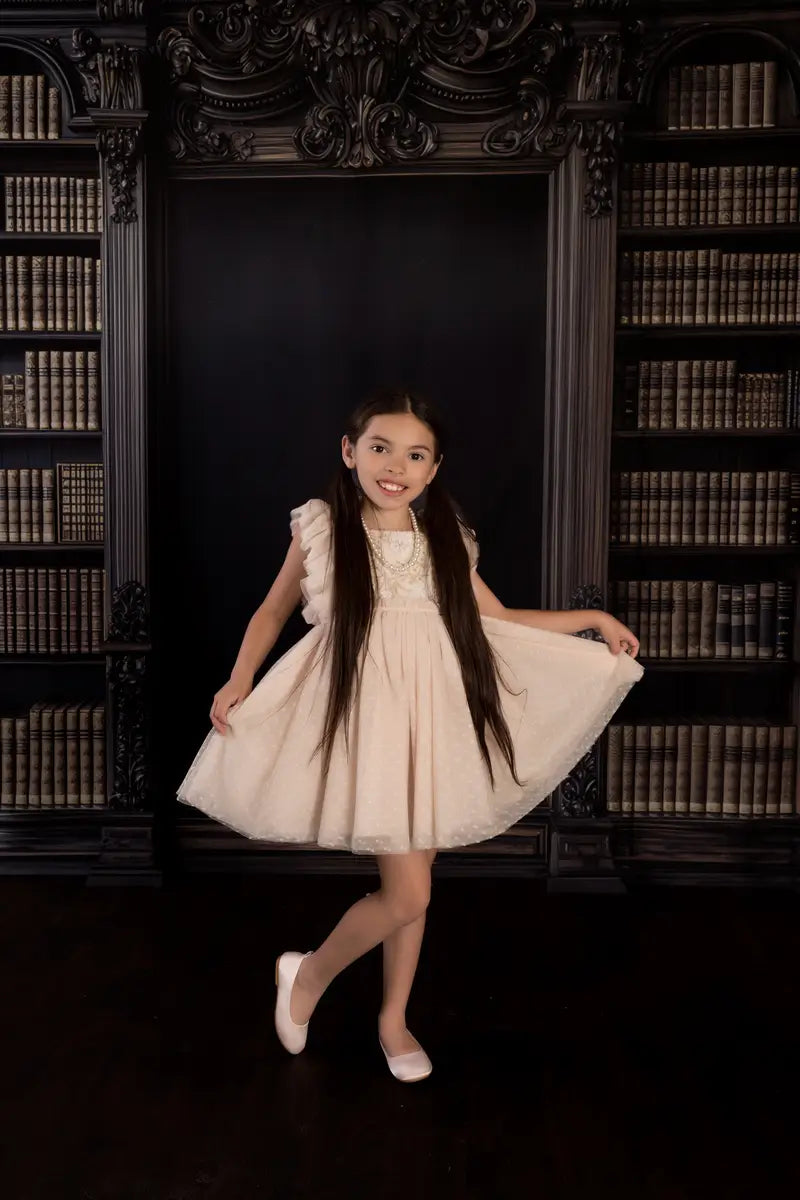 A young girl in a beige dress posing in front of a vintage library chalkboard graduation backdrop with an academic bookshelf background.