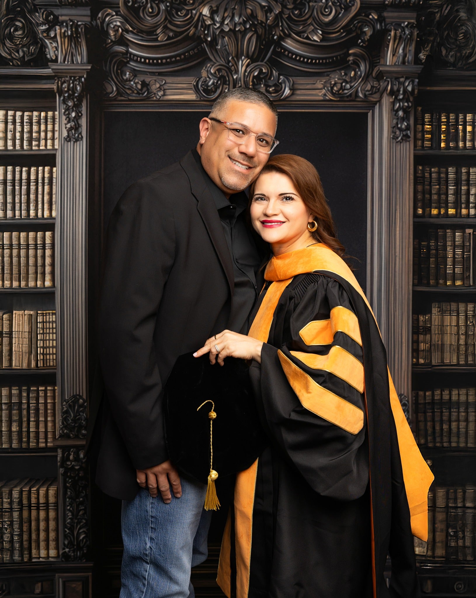 A middle-aged woman holding a diploma and posing with her husband in front of a vintage library chalkboard graduation backdrop with an academic bookshelf background.