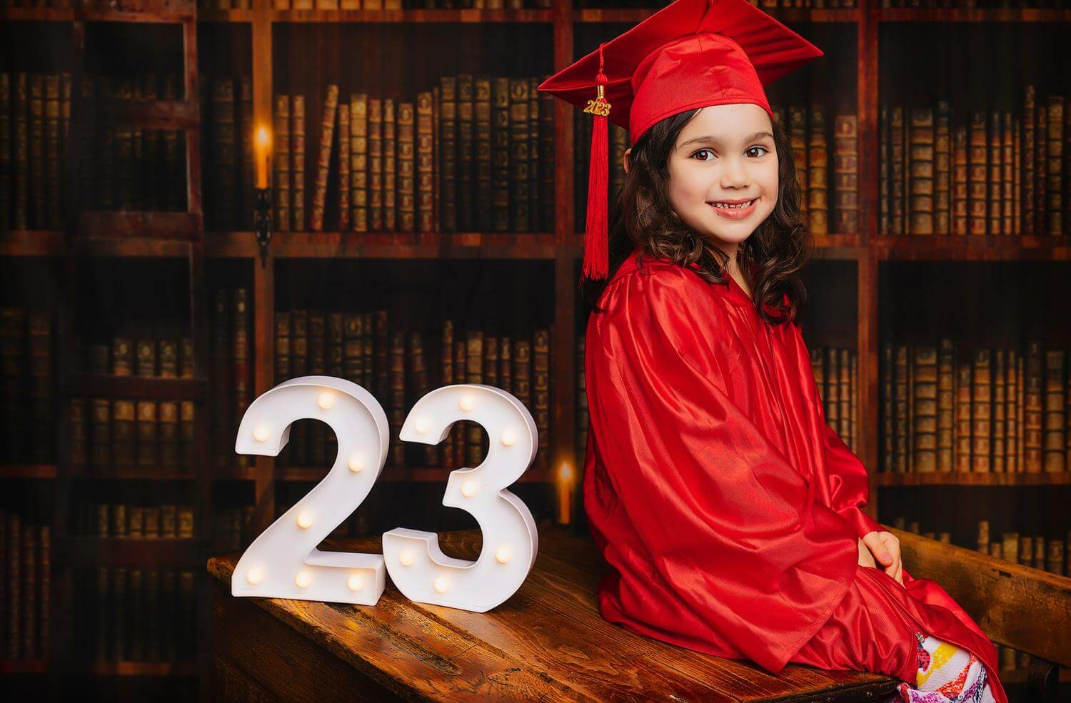 A little girl wearing a red graduation gown sitting on a table in front of a vintage library photo background with decorative number 23 on the table.