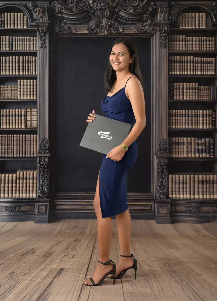 A female college graduate holding a diploma while standing in front of a vintage library chalkboard backdrop with an academic bookshelf background.