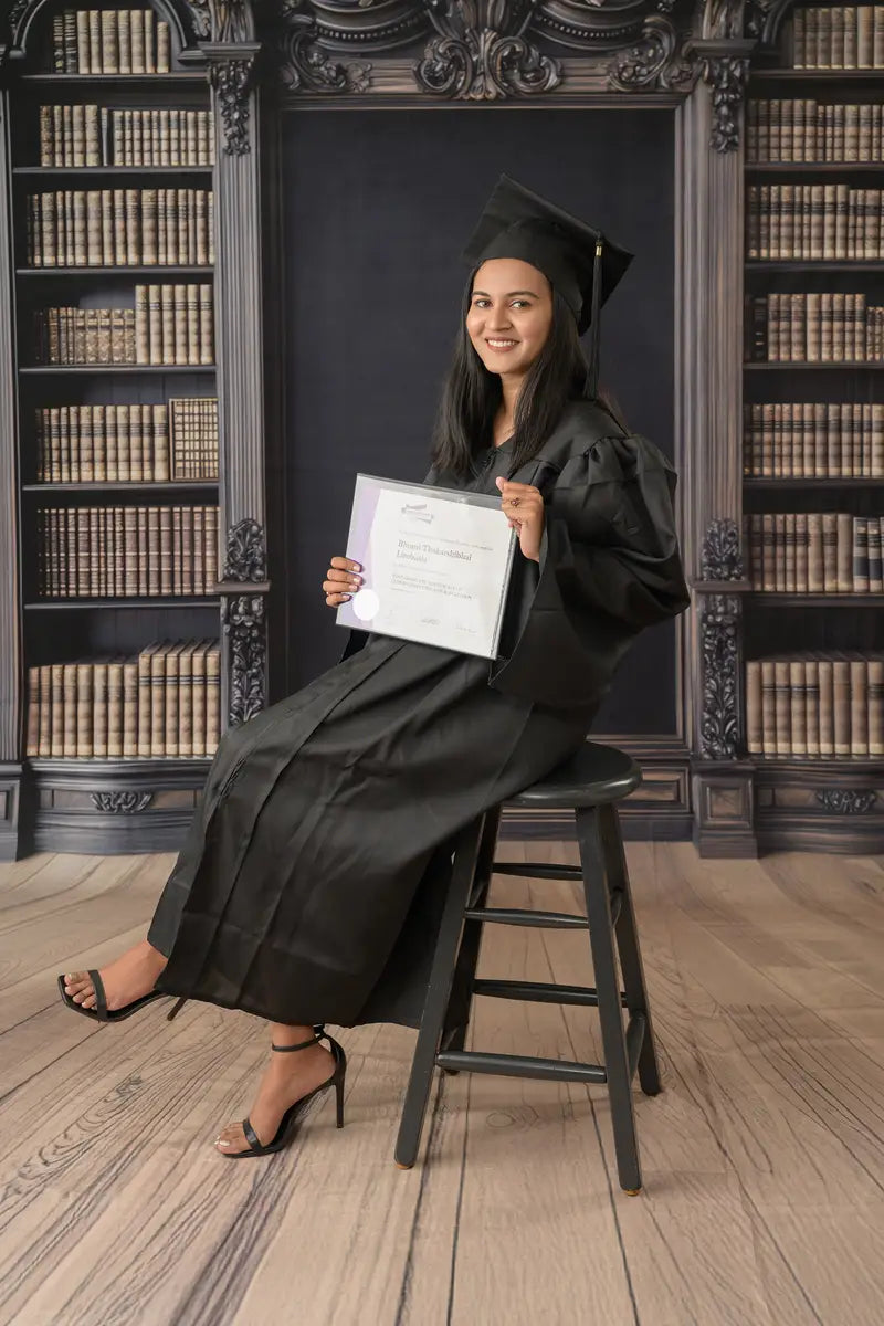 Graduate in black cap and gown holding a diploma, sitting on a stool in front of a bookshelve photo backdrop.