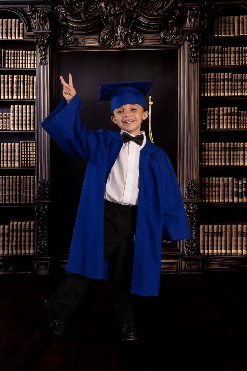 An elementary school student making a peace sign while wearing a blue graduation gown in front of an academic bookshelf photo background.