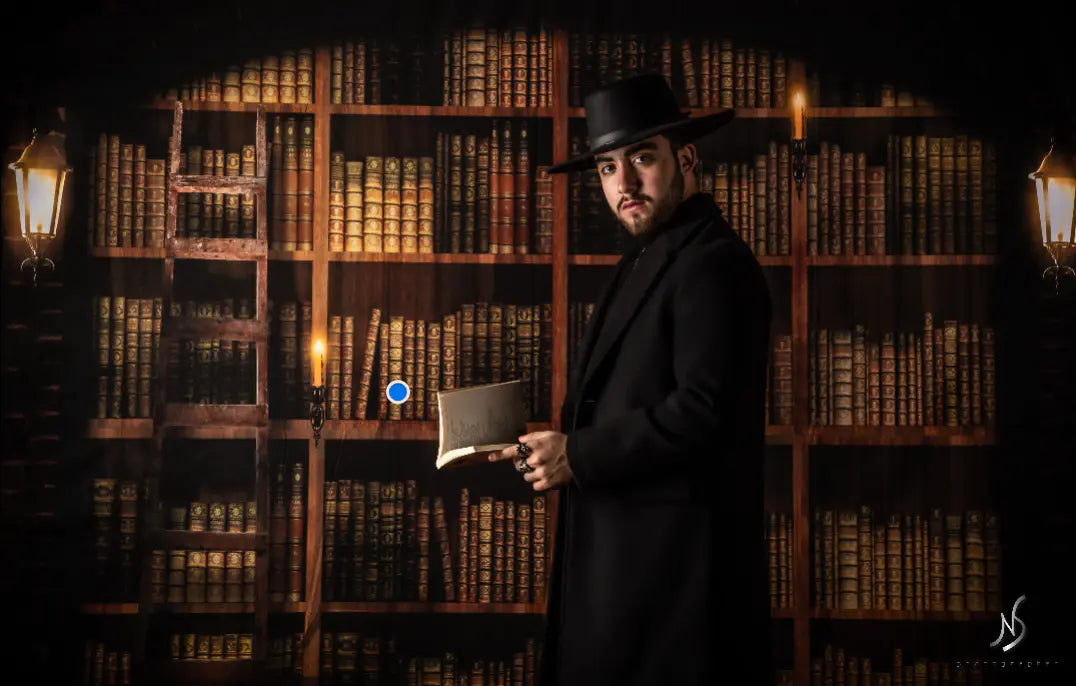 Man in a dark suit and hat holding a book in front of a bookshelf photography backdrop.