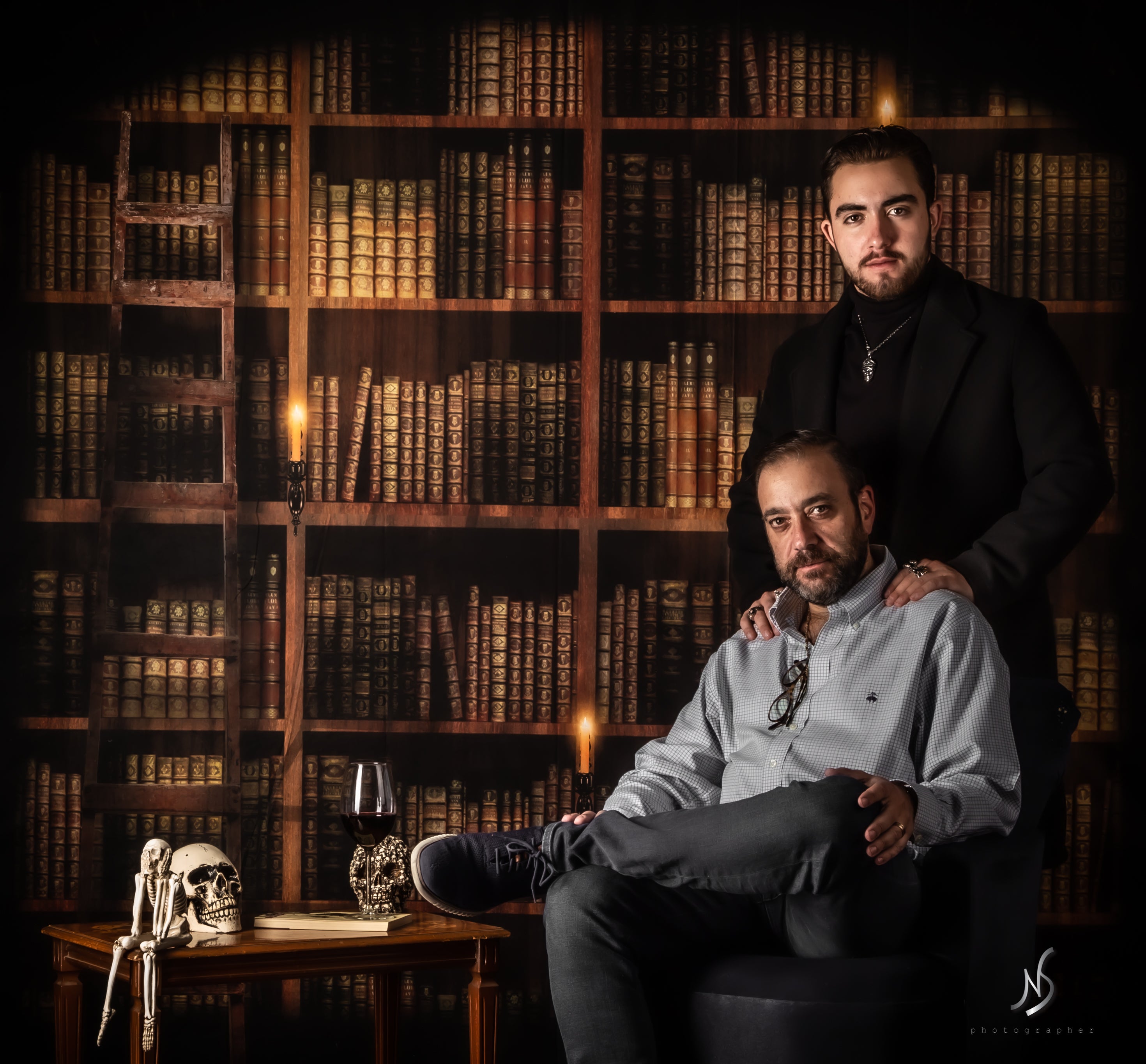 Two male friends posing for a graduation photo in front of a vintage library bookshelf backdrop, one seated and the other standing behind with hands on his shoulders, with a skull decoration on the table.