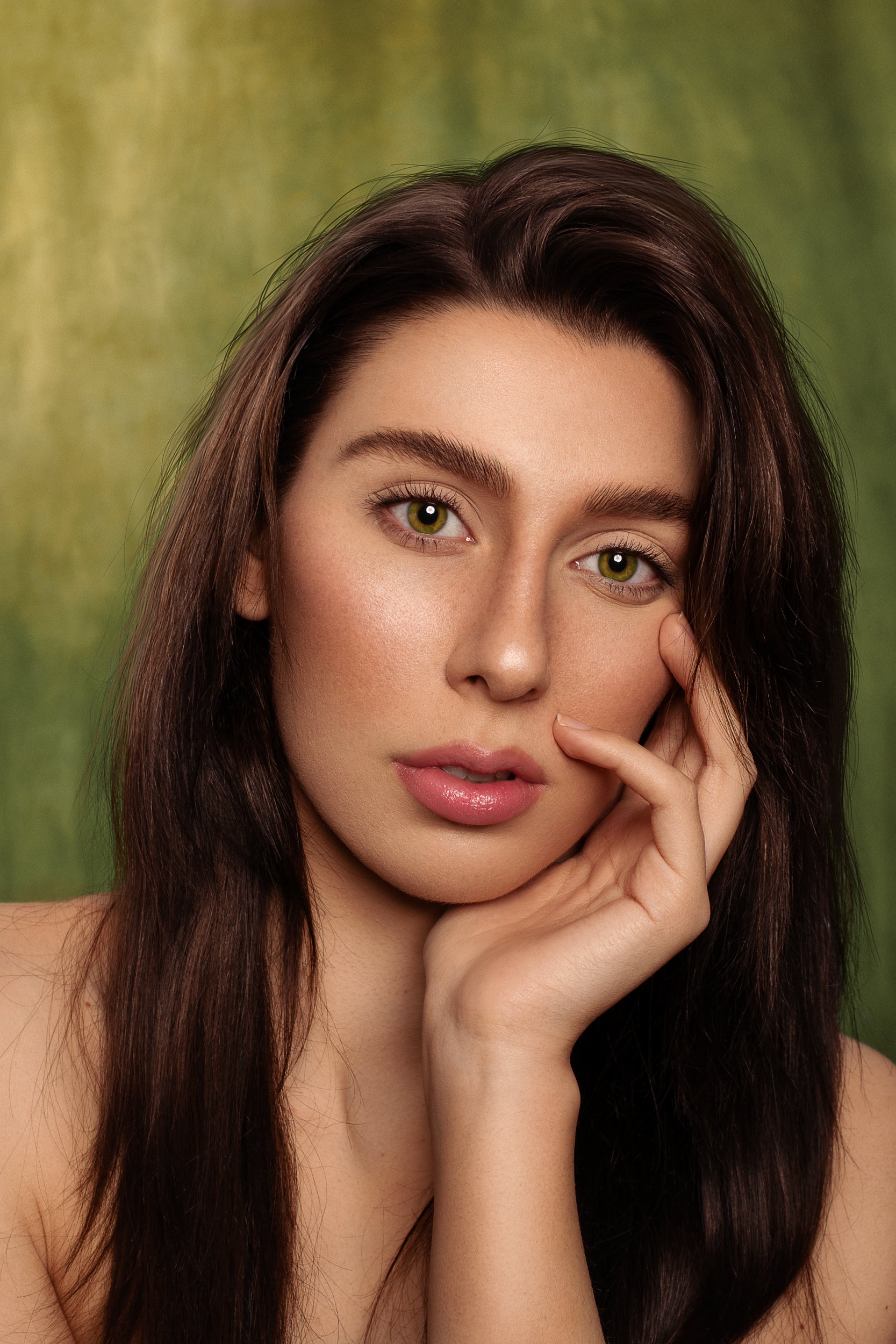Portrait of a woman with long brown hair and striking green eyes, gently touching her face against a textured backdrop of Kate Sweep Green Abstract Backdrop for Photography. She has natural makeup and a serene expression.
