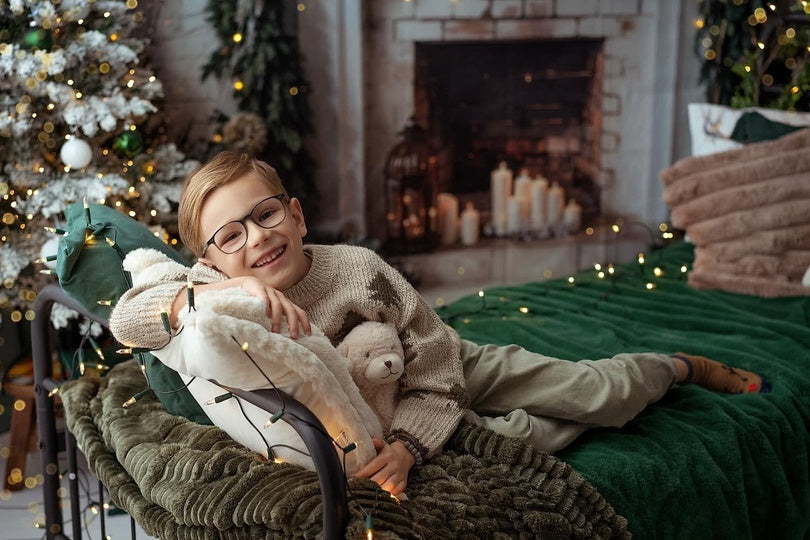 Young boy relaxing in a cozy winter portrait scene with soft blankets, warm lights, a fireplace, and festive holiday details.