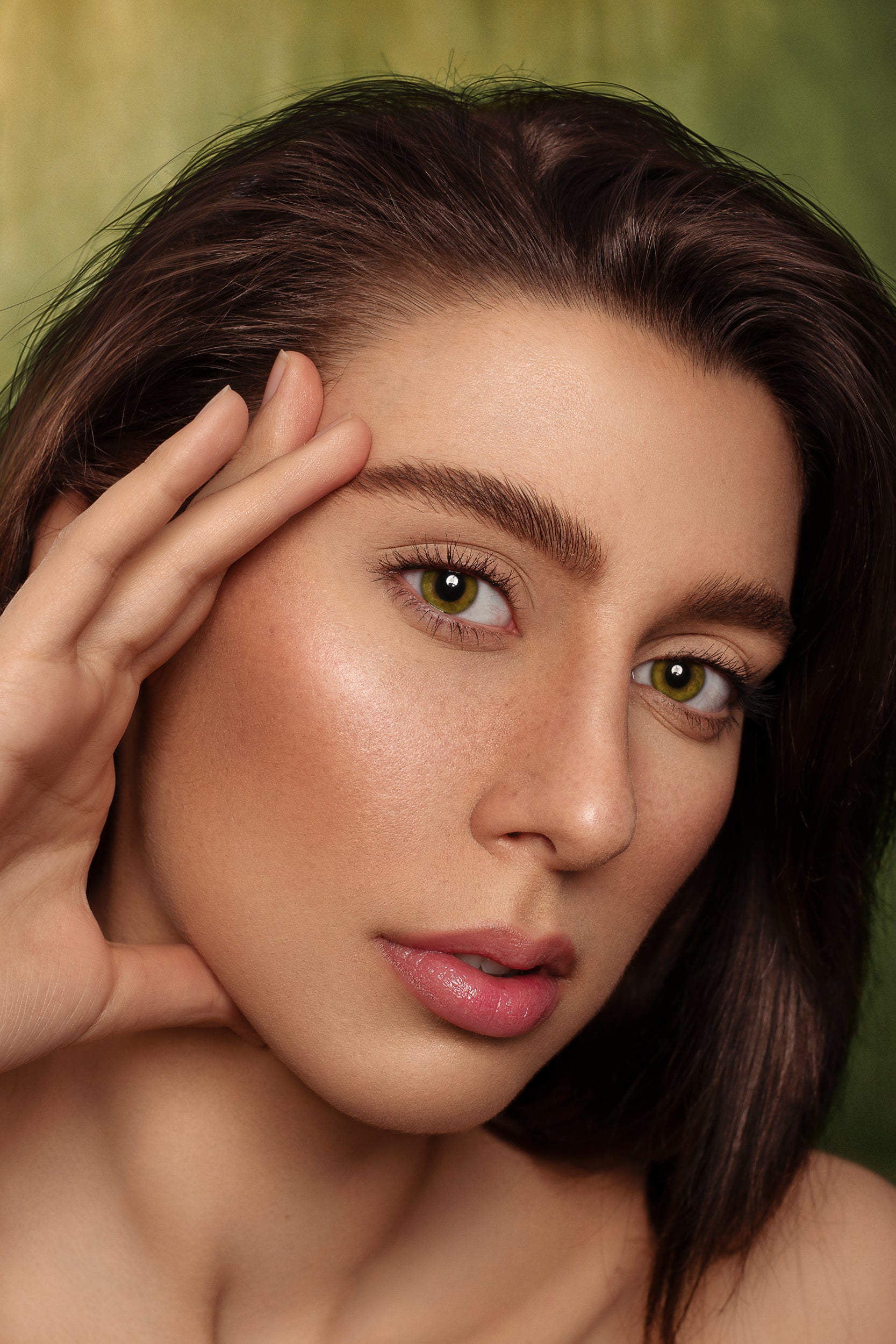 Close-up portrait of a young woman with brown hair and striking green eyes, lightly touching her face against the Kate Sweep Green Abstract Backdrop for Photography. Her makeup is subtle yet enhances her natural features.