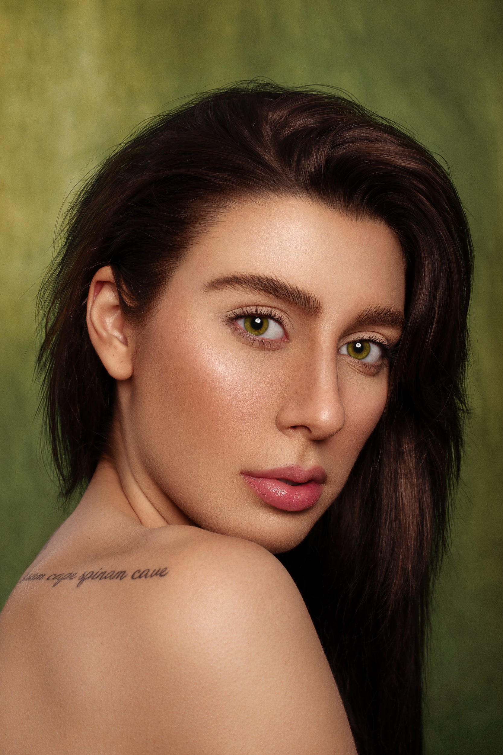 Portrait of a woman with long dark hair and green eyes, looking over her shoulder against a Kate Sweep Green Abstract Backdrop for Photography. She has subtle makeup and a visible tattoo on her shoulder.