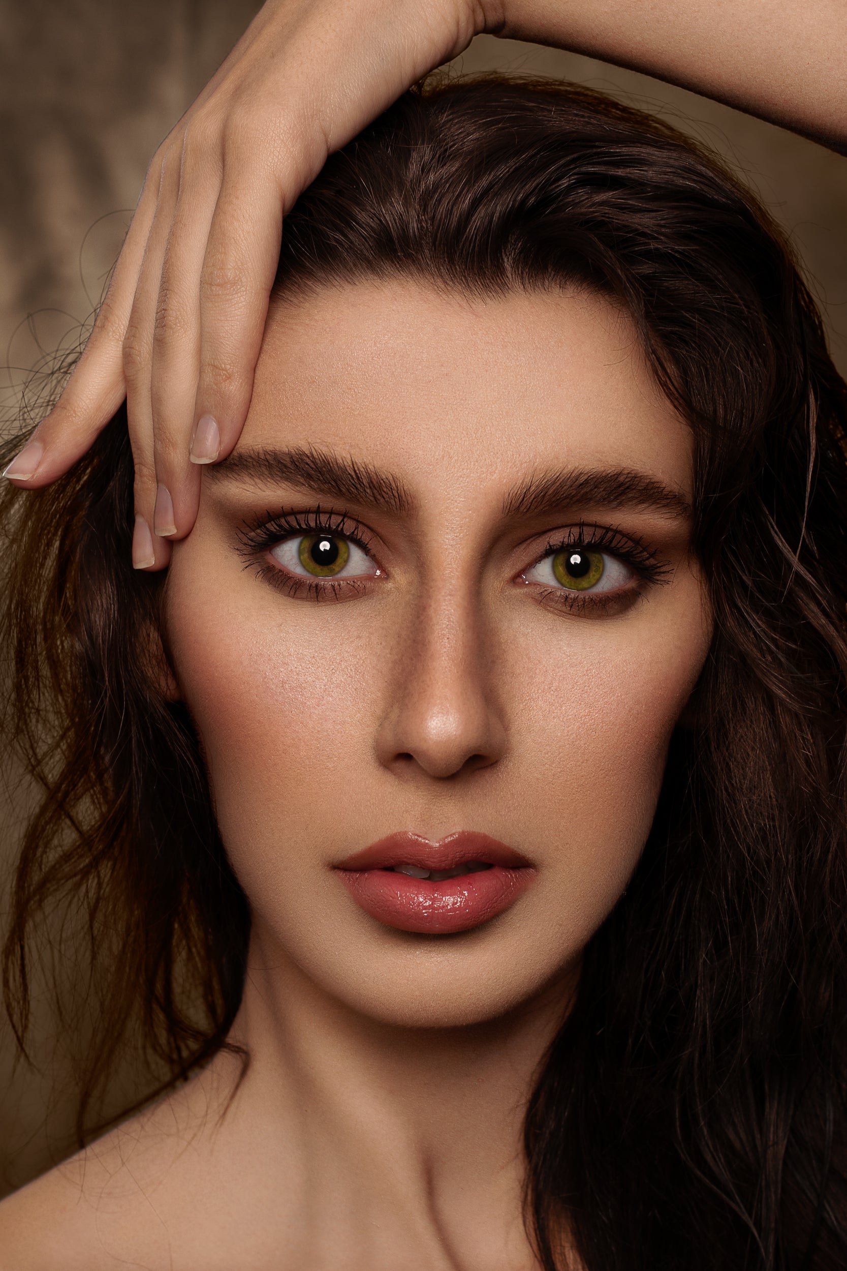 Close-up portrait of a woman with intense green eyes, glossy lips, and wavy brown hair using Kate Sweep Green Abstract Backdrop for Photography, her hand gracefully touching her forehead. Her expression is captivating and serene.